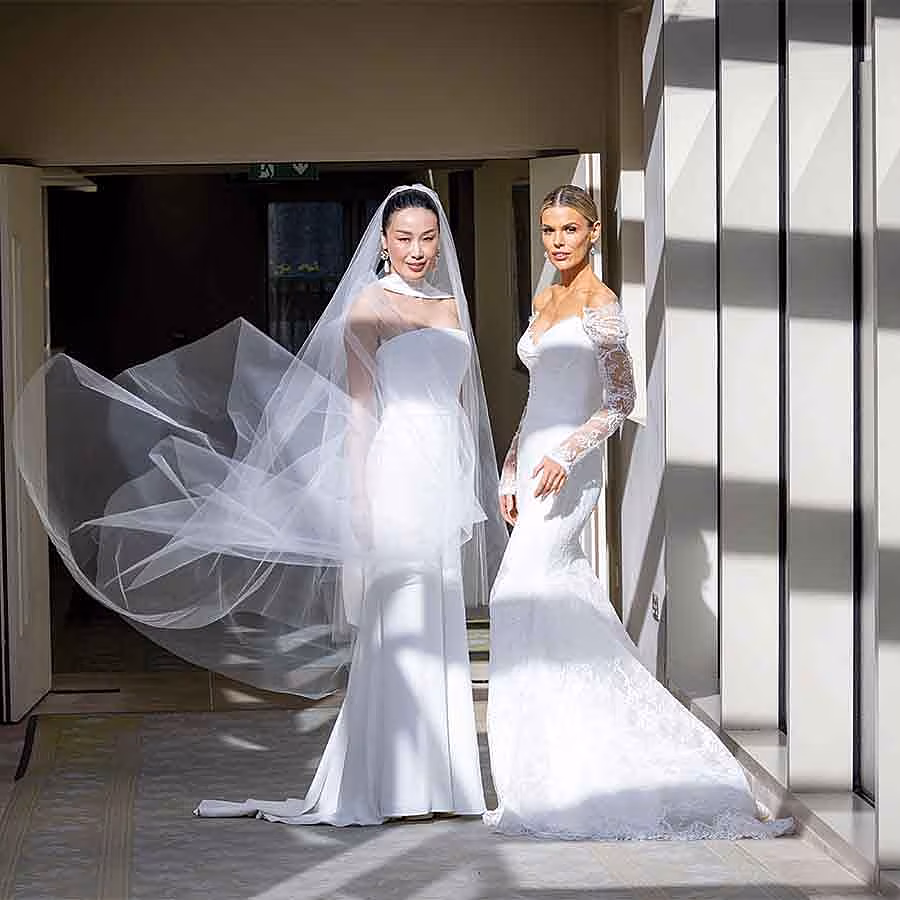 Two brides wearing elegant white wedding gowns stand in a sunlit hallway, one with a flowing veil and the other in a lace-sleeved dress.